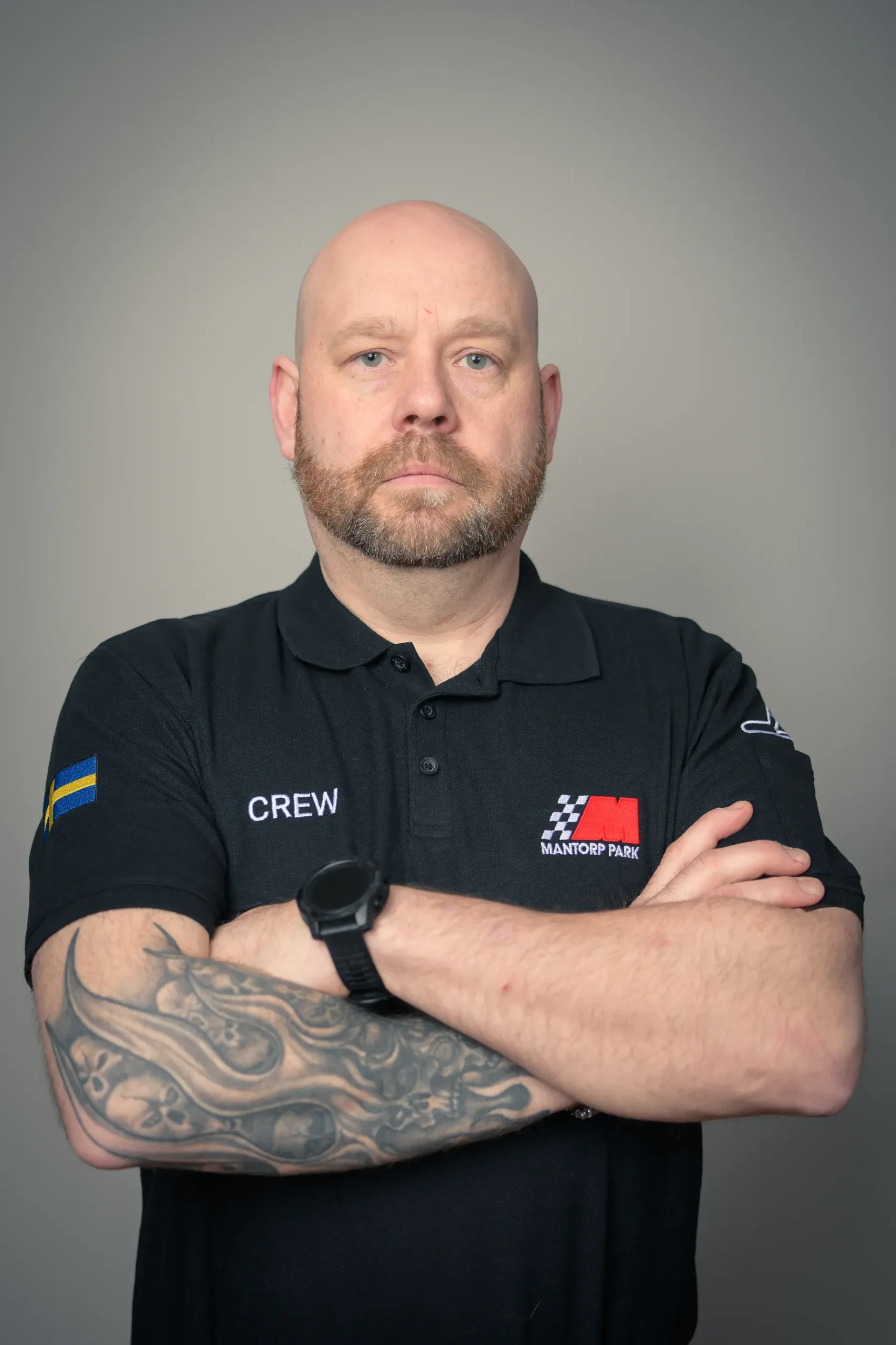 En skallig man med tatueringar på vänster arm står med armarna i kors. Han är klädd i en svart pikétröja med CREW och en logga där det står Mantorp Park. Det finns en svensk flagga på ärmen. Bakgrunden är vanlig grå.
