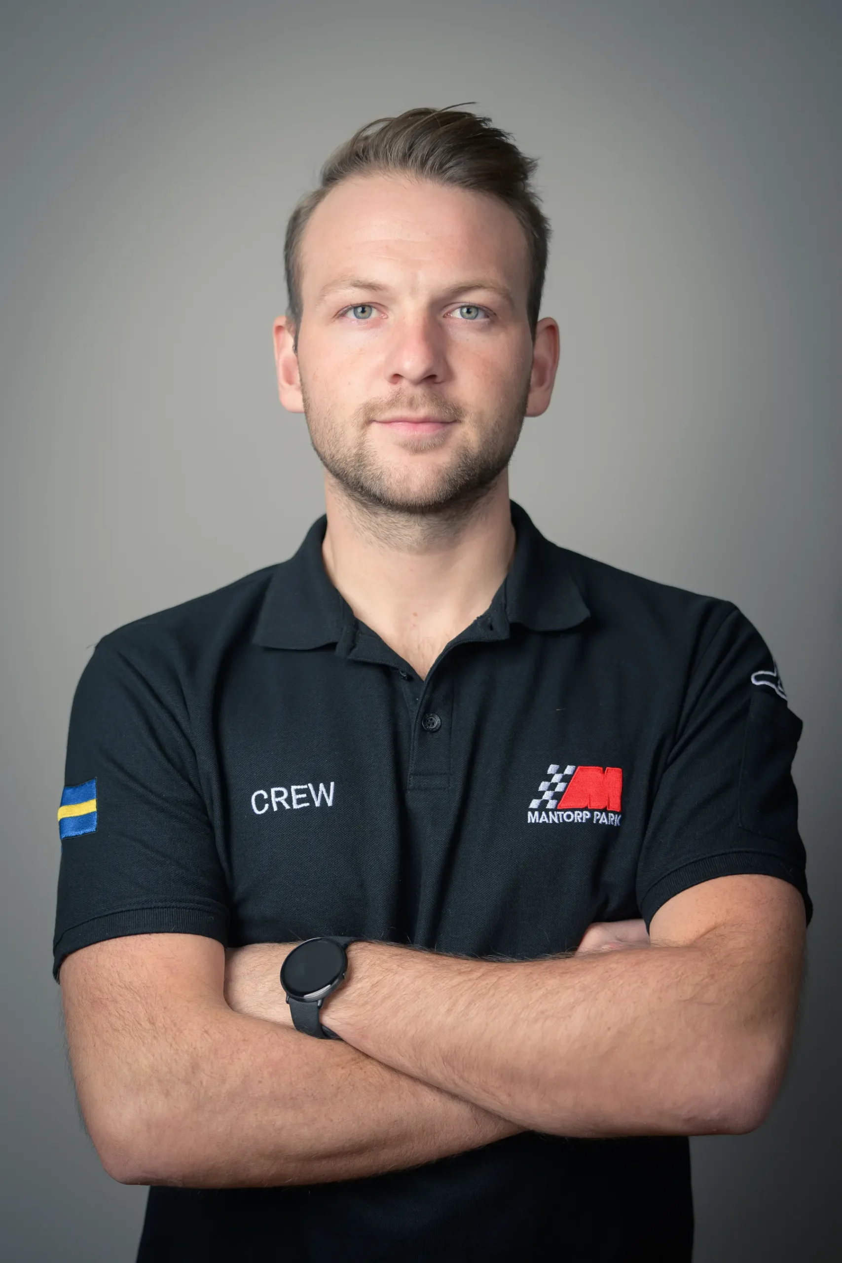 En man med kort hår och skägg står med armarna i kors, iklädd en svart pikétröja med logotyper och ordet CREW. Han har ett neutralt uttryck. Bakgrunden är en enkel grå gradient.