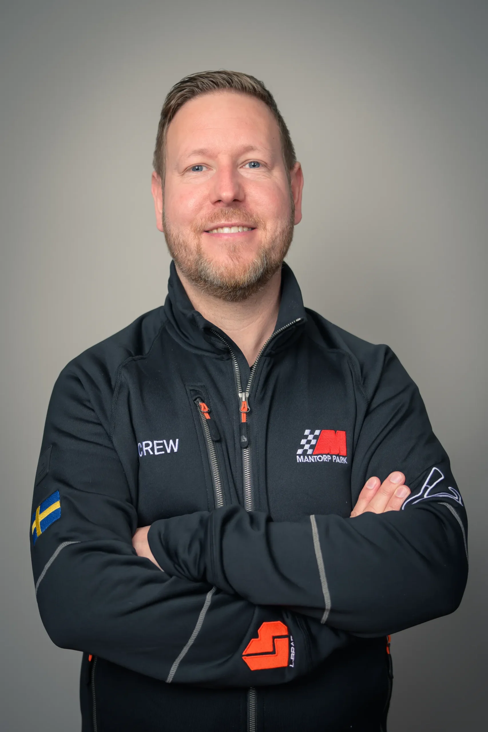 En person med kort hår och skägg ler, iklädd en svart jacka med racingbrickor och en svensk flagga på ärmen. Deras armar är korsade. Bakgrunden är vanlig grå.