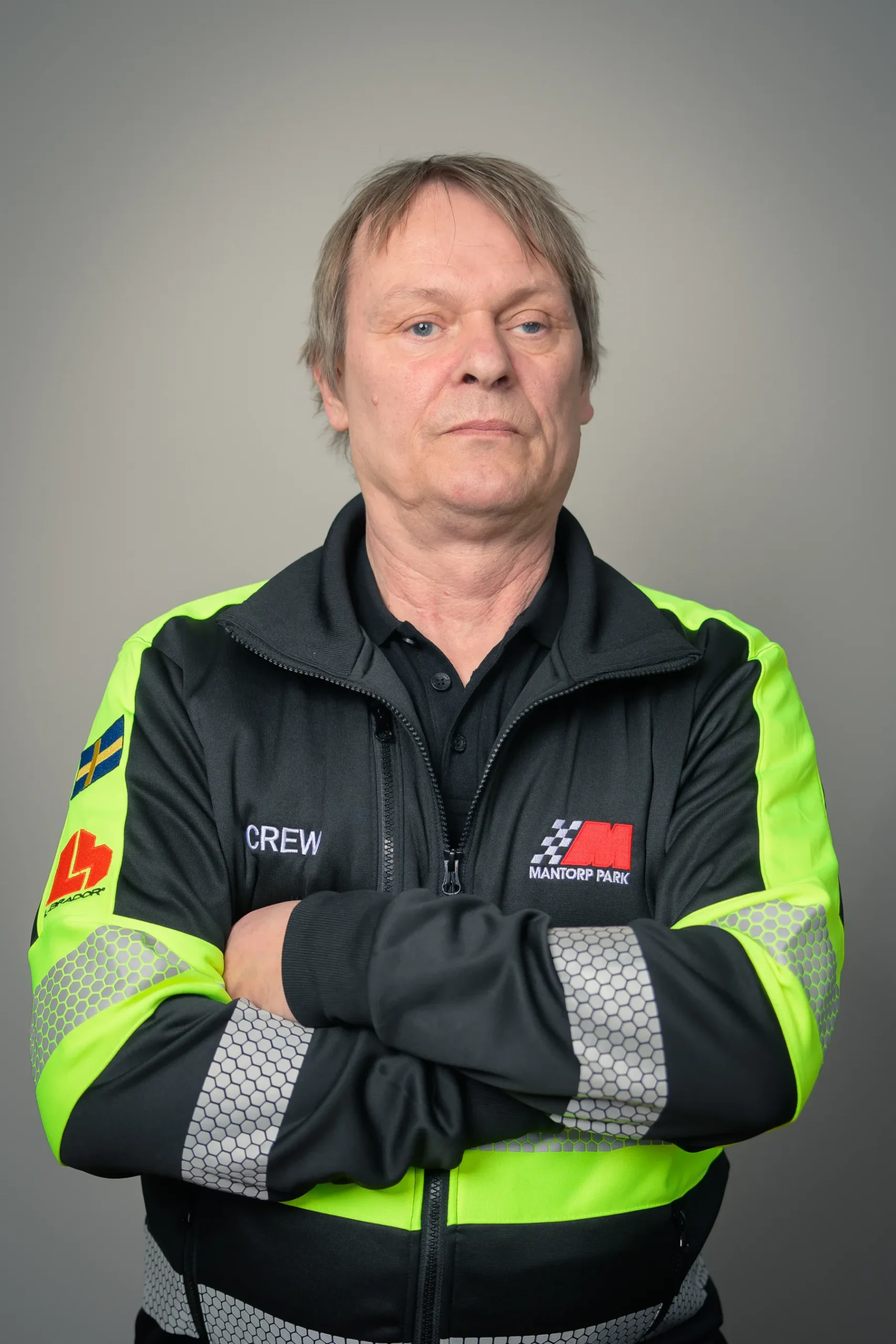 En person med kort hår korsar armarna och bär en synlig jacka med CREW och flera logotyper. Jackan har ljusgula partier och är inställd mot en neutral grå bakgrund.