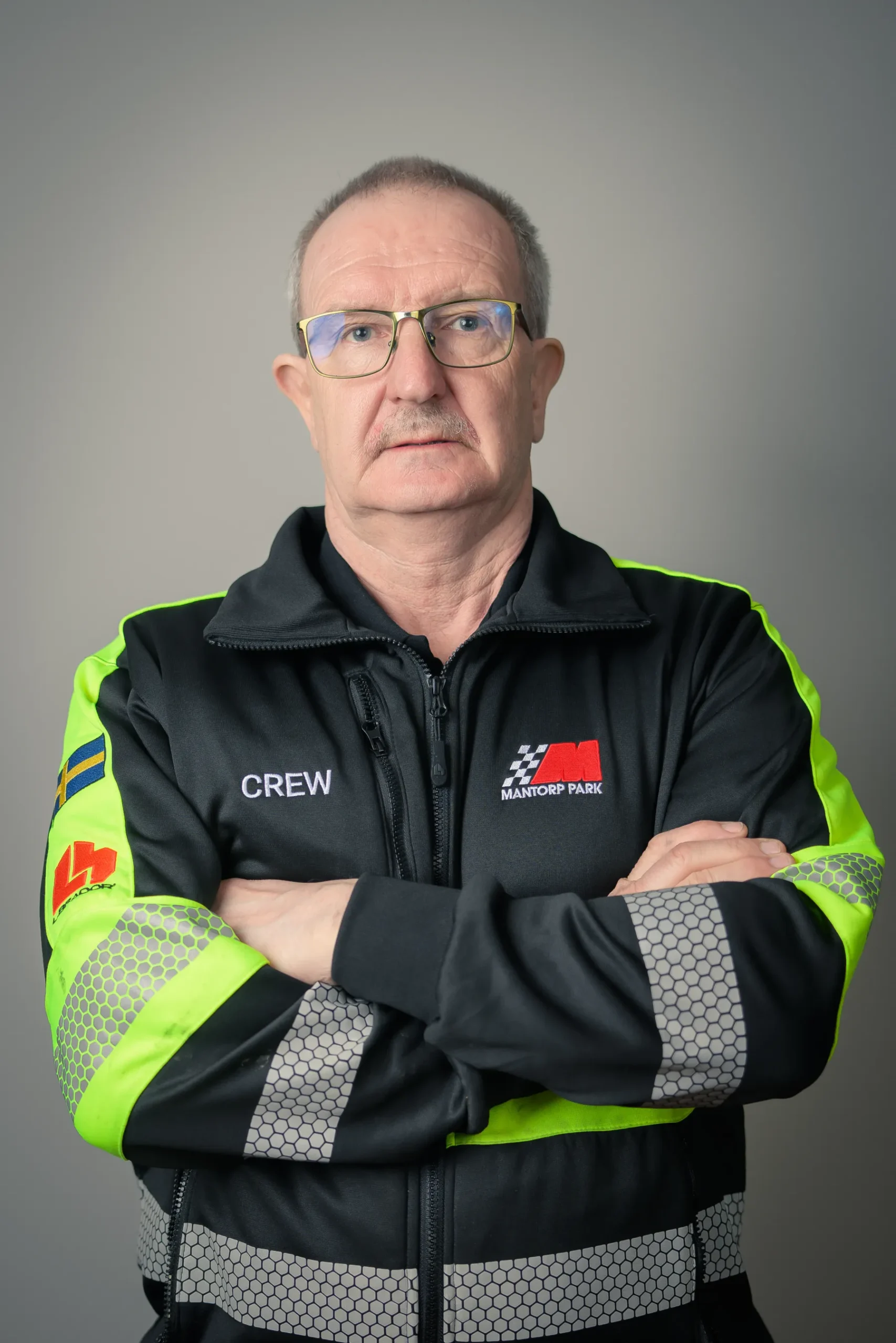 Man med kort hår och glasögon, klädd i en svart och neongrön jacka med CREW skrivet på. Jackan har olika patchar. Han står med armarna i kors mot en neutral bakgrund och tittar direkt in i kameran.