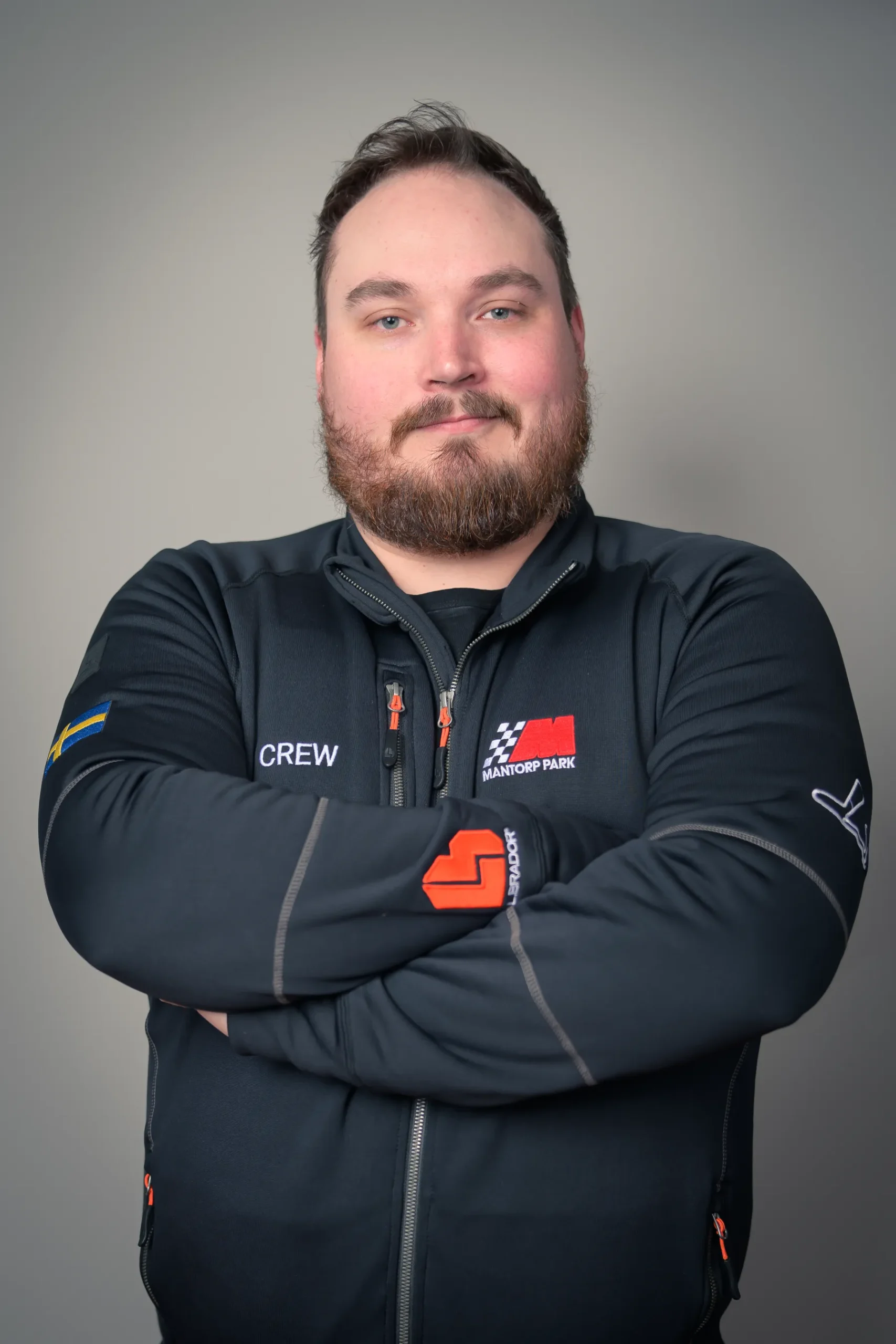 En skäggig individ står med armarna i kors, iklädd en svart jacka med logotyper och ordet CREW på. Personen har ett neutralt uttryck och ställs mot en vanlig grå bakgrund.