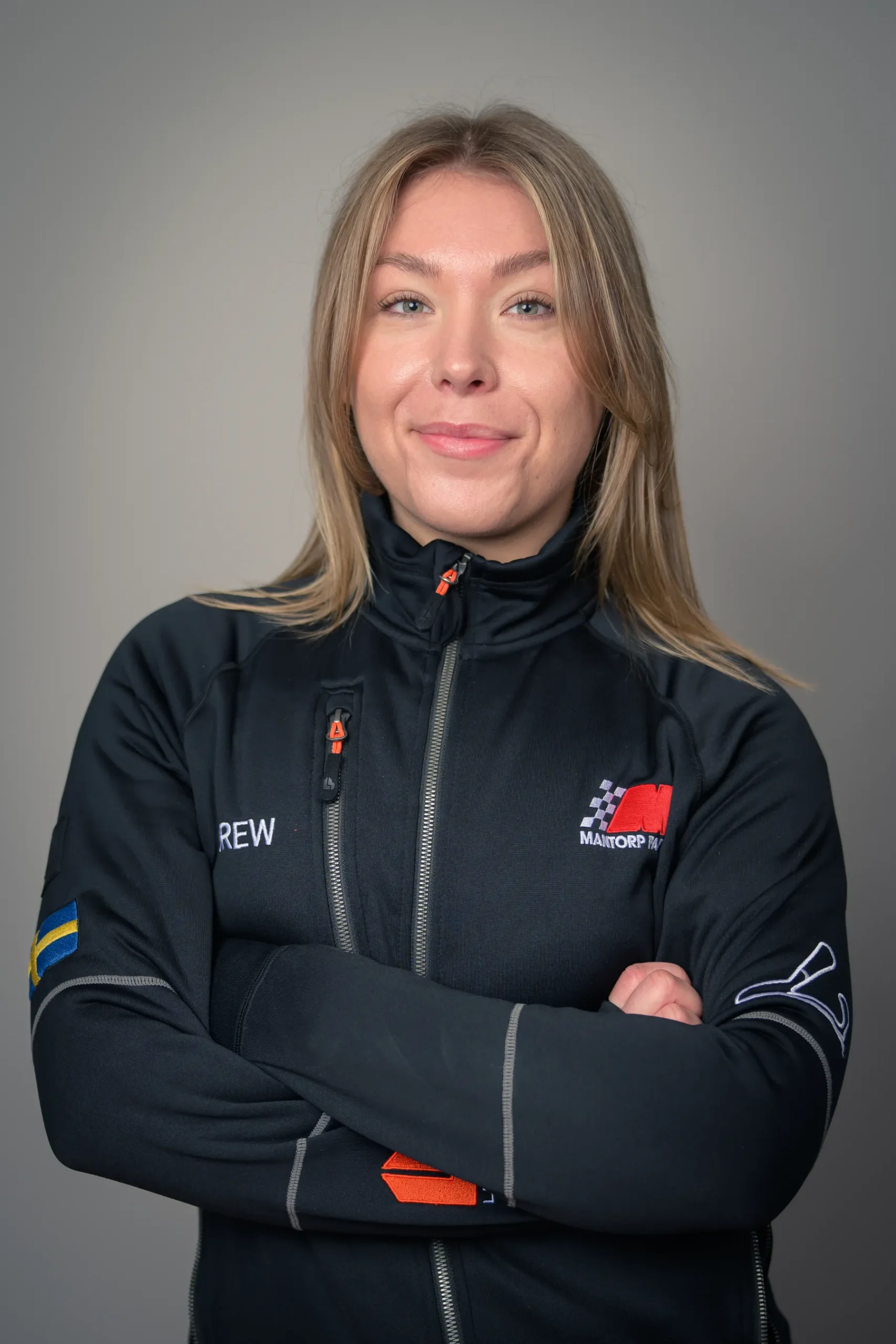 En person med långt blont hår står självsäkert med armarna i kors, iklädd en svart jacka med flagglapp och logotyper. Bakgrunden är neutral och grå, vilket framhäver personen i mitten.