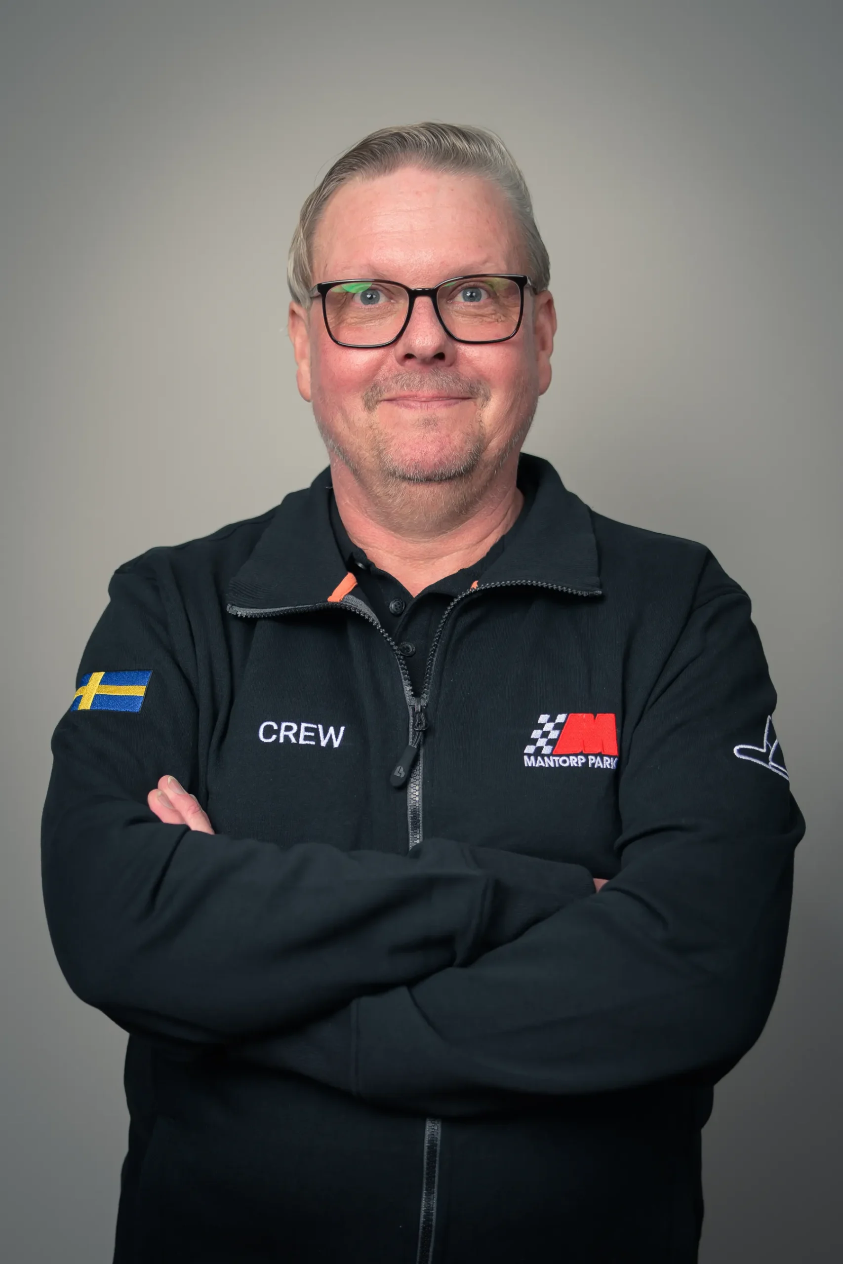 En man står med armarna i kors, iförd glasögon och en svart jacka med en svensk flagga-lapp och ordet CREW på. Jackan har även en logotyp för Mantorp Park, en racingbana. Bakgrunden är neutral.