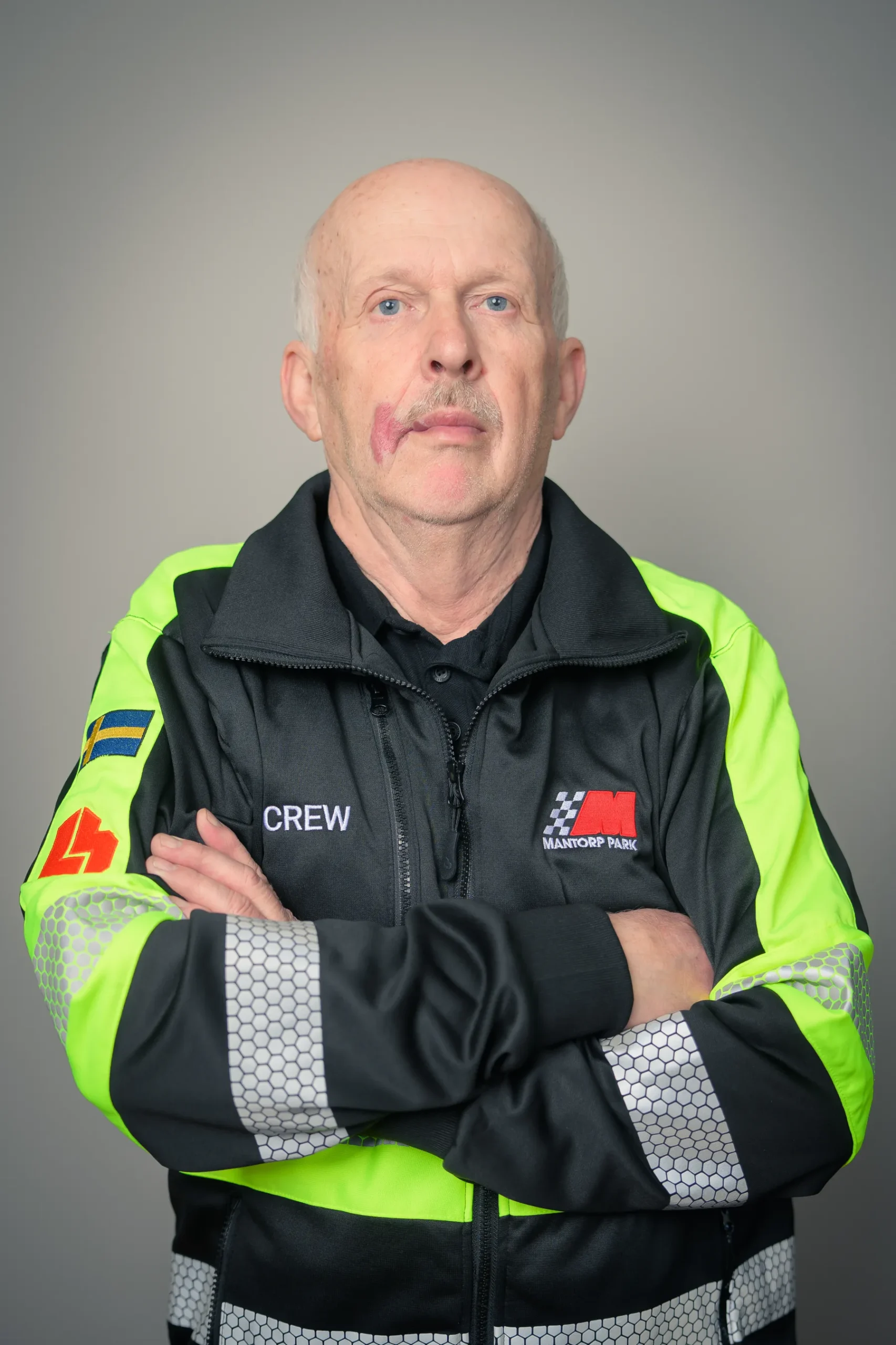 En äldre man står med armarna i kors iförd en svart och neongrön säkerhetsjacka med reflekterande ränder. Jackan har en logotyp och CREW tryckt på. Han har kort hår och ett synligt födelsemärke på kinden, poserar mot en grå bakgrund.