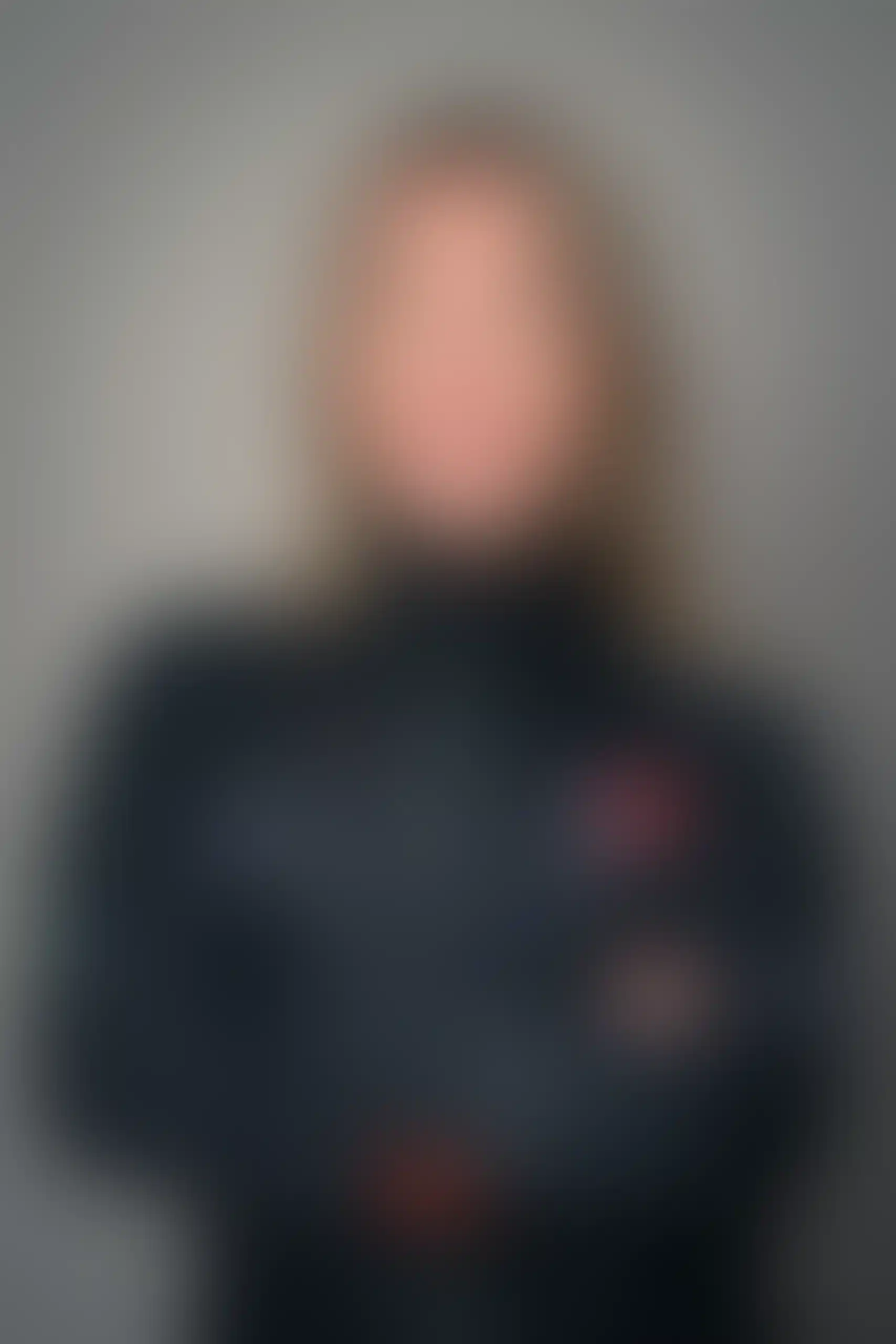 Suddig bild av en person med långt hår som bär en mörk jacka, armarna i kors, mot en vanlig bakgrund. Ansiktsdragen och detaljerna är otydliga på grund av suddigheten.