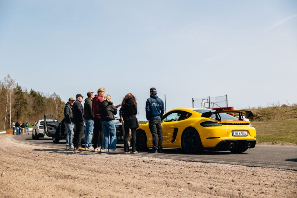En grupp människor står vid en gul sportbil som är parkerad på en bana, med en annan bil i närheten. Miljön är utomhus en klar dag, med några träd och gräsytor i bakgrunden.