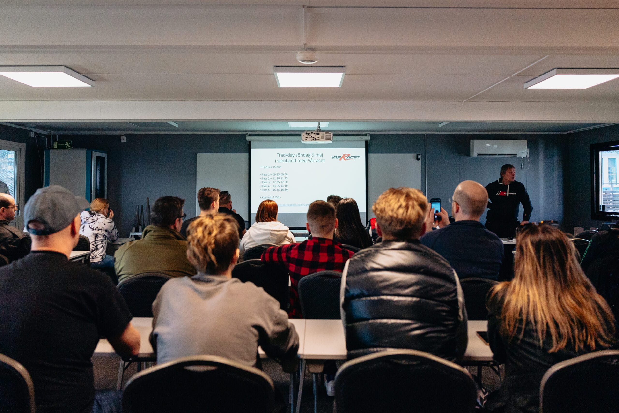 En grupp människor sitter i ett klassrum och tittar på en skärm där en man står längst fram och håller en presentation. På skärmen visas text och en logotyp, och deltagarna tittar uppmärksamt på eller tar foton.