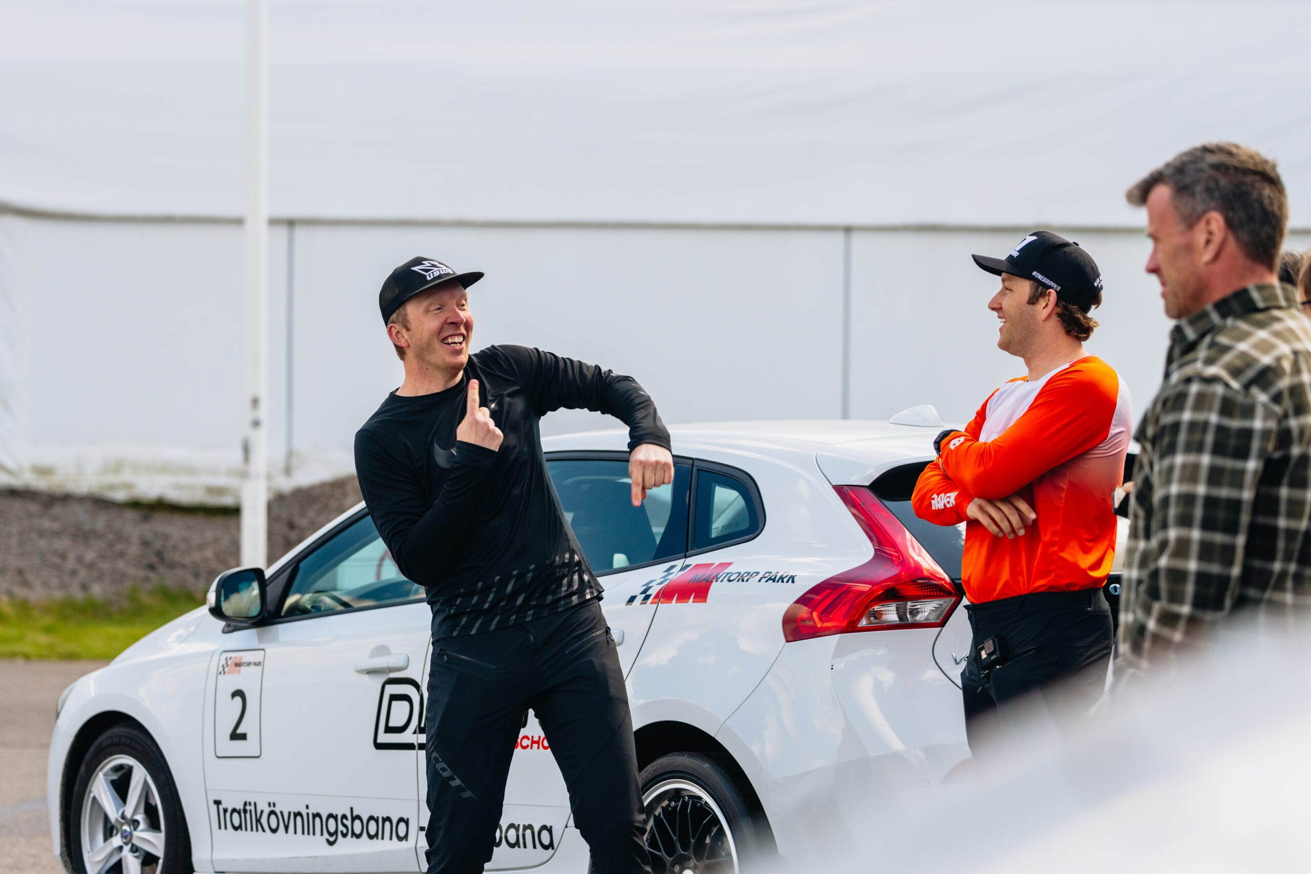 Tre personer står bredvid en vit bil med racingdekaler. En man gestikulerar livligt och ler, medan de andra tittar och lyssnar. De verkar ha en livlig konversation utomhus i närheten av en racerbana.