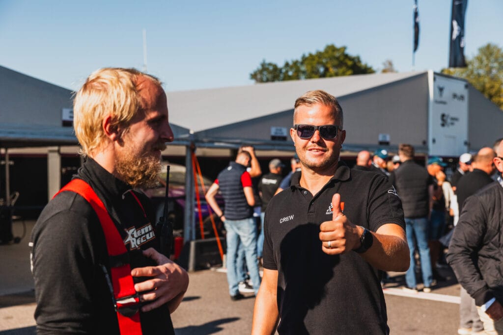 Två män står utomhus på ett evenemang; den ene gör tummen upp och bär solglasögon, medan den andre ler med en hand på bröstet. Människor och tält syns i bakgrunden under en klar himmel.