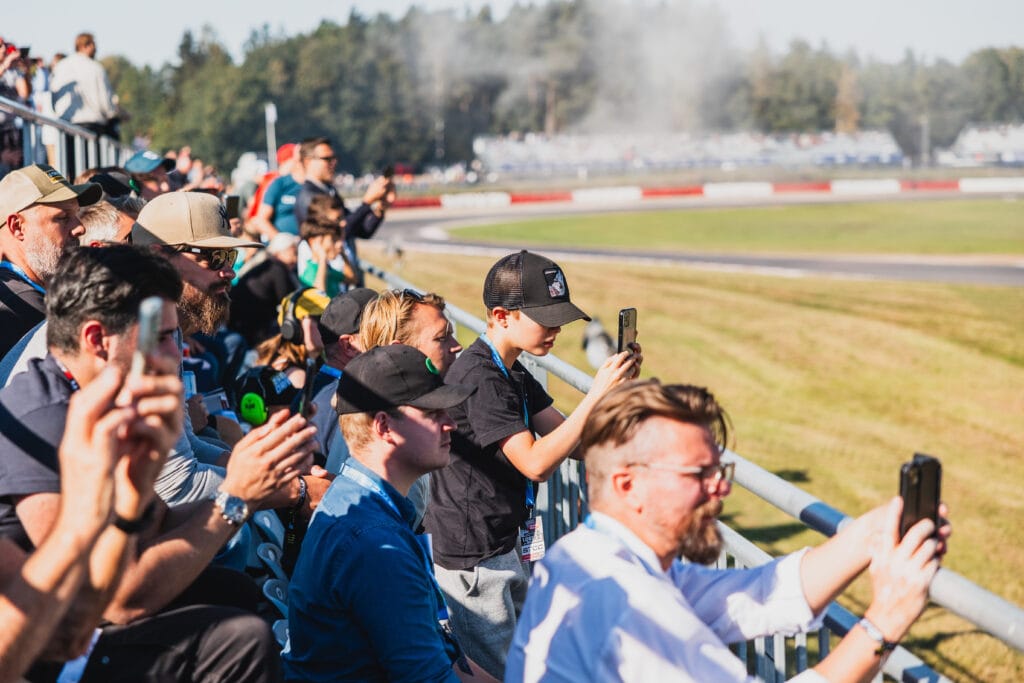 En folkmassa i stadionstolar tittar på ett bilrace, många håller upp telefoner för att ta foton eller videor. Banan och ett rökmoln syns i bakgrunden under en solig himmel.