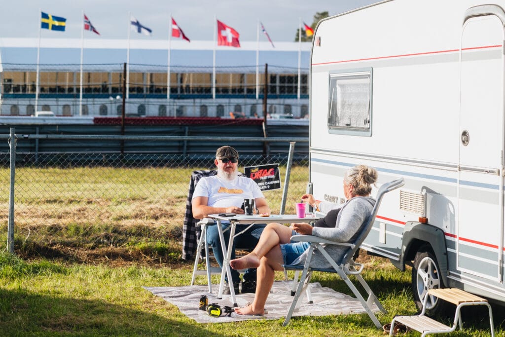 Två personer kopplar av utanför en husvagn, sittande på campingstolar vid ett litet bord på en gräsyta. Flera nationalflaggor vajar i bakgrunden nära en kapplöpningsbana eller en evenemangsplats.