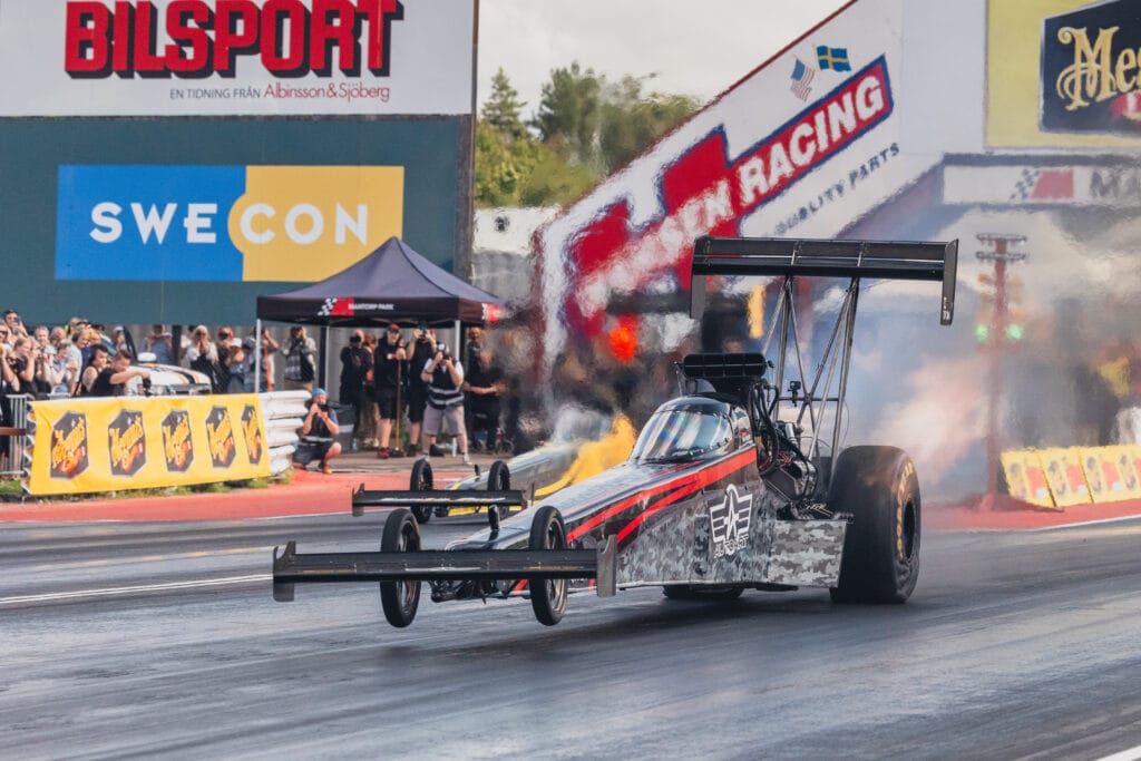 En dragracingbil accelererar nedför en bana med rök från däcken, med en publik och sponsorbanderoller i bakgrunden.