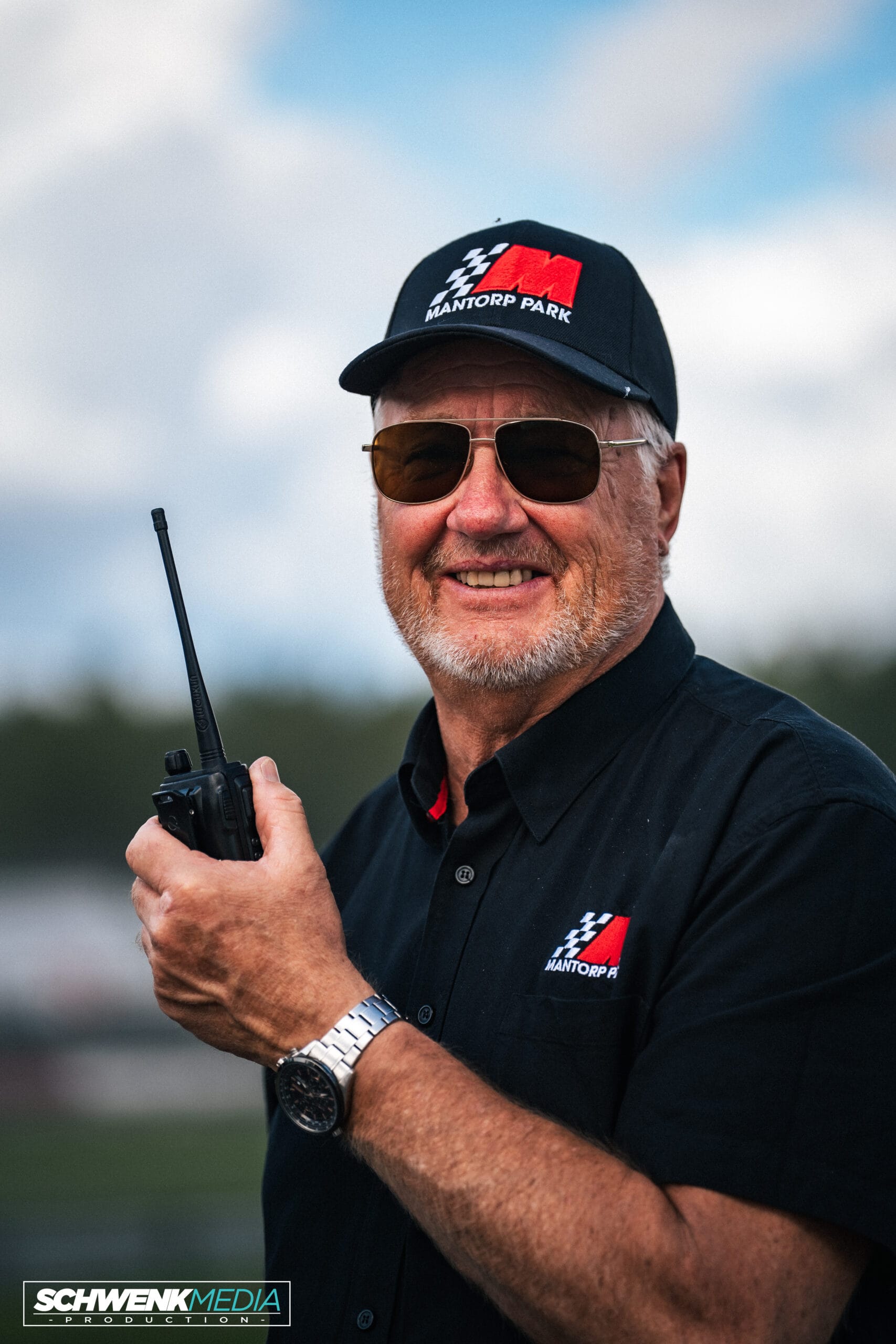 En man i solglasögon och svart keps och skjorta med "Mantorp Park"-logotyper ler medan han håller i en walkie-talkie utomhus. Bakgrunden är suddig och visar blå himmel och grönska.