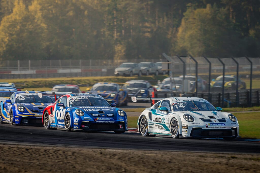 Flera racerbilar kör i hög fart runt en kurva på en racerbana, med den ledande bilen i vitt och de andra tätt efter. Träd, stängsel och suddiga fordon syns i bakgrunden i dagsljus.