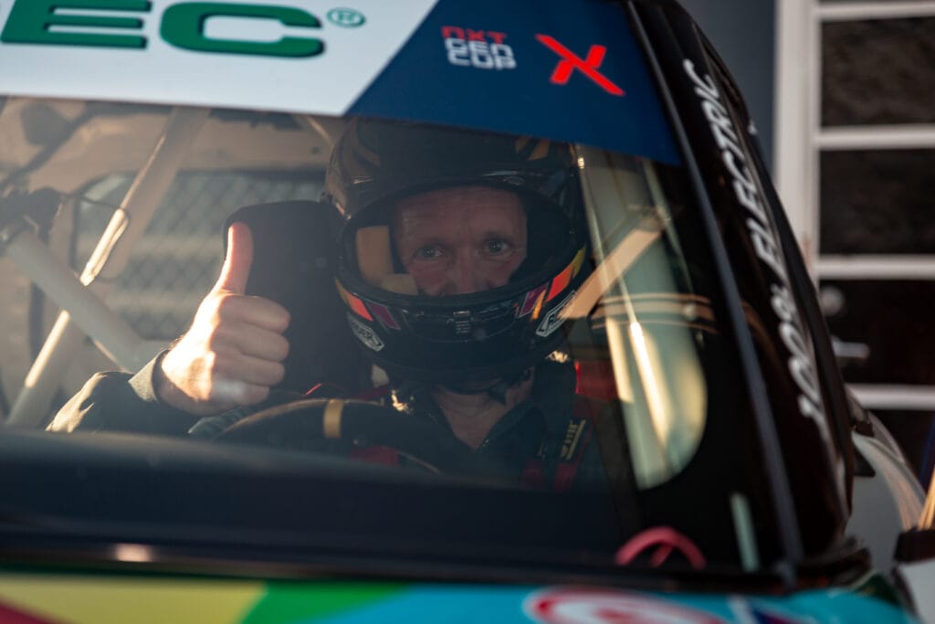 En racerbilsförare med hjälm sitter i en bil och gör en tummen upp-gest genom fönstret, vilket tyder på godkännande eller beredskap inför ett lopp.