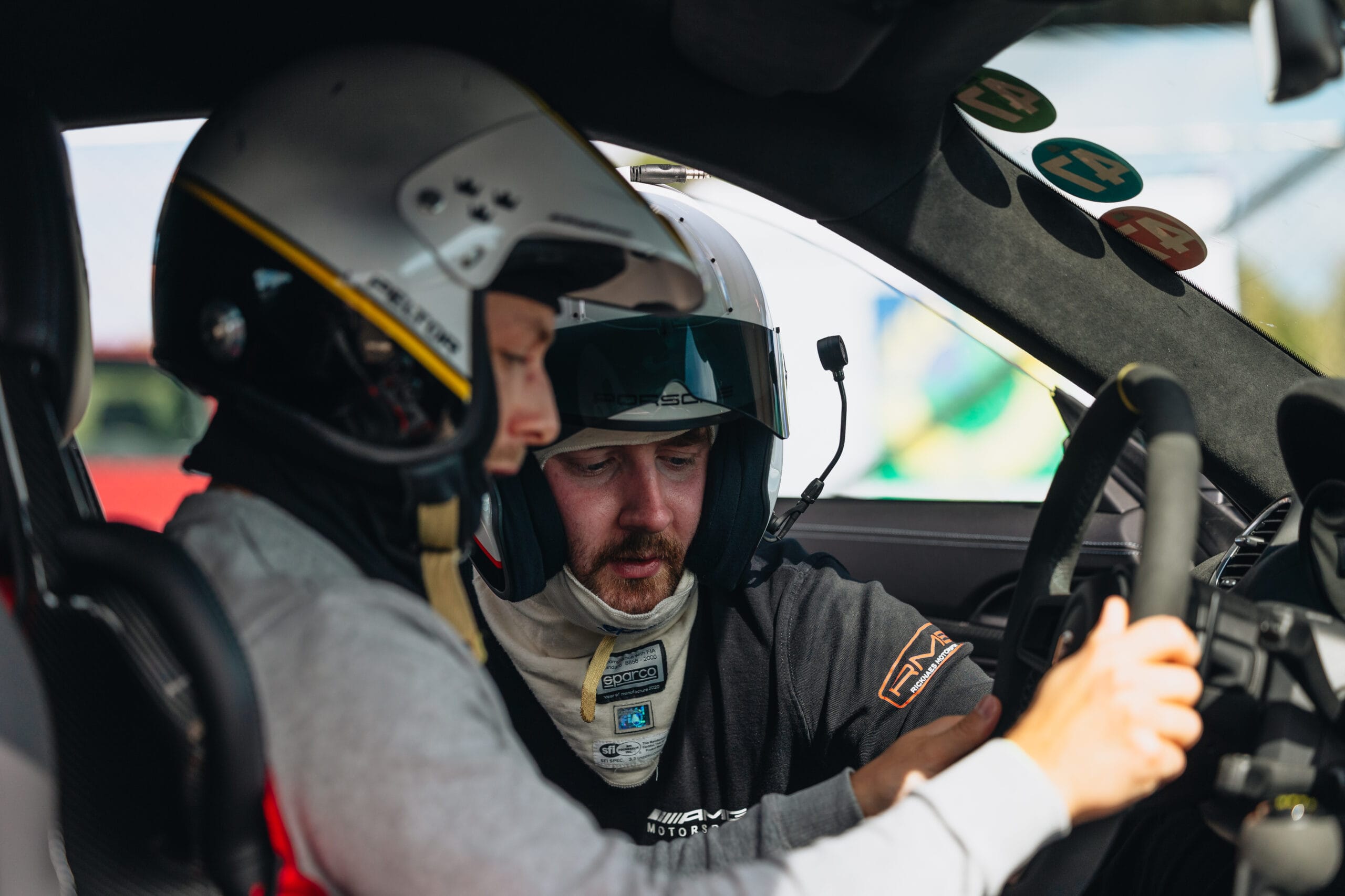 Två personer med racinghjälmar på sig sitter i en bil. En person sitter i förarsätet och håller i ratten, medan den andra lutar sig in från passagerarsidan och ger instruktioner eller diskuterar något intensivt.